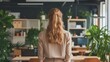 © fara - Woman with long hair, back view, standing in a modern, plant-filled office.