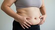 © SimpleDesignStudio - A close-up of a woman's midsection, showcasing her stomach, revealing a healthy yet round belly. She is standing and appears to be assessing her body positively, in a bright indoor setting.