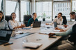 © qunica.com - A group of multicultural business people engaged in a meeting at a large conference table. They are discussing ideas with laptops and documents in a modern office environment.