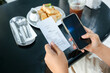 © patpitchaya - Closeup hands of a woman holding smart phone and food receipt, with toast and a cold drink on the table in the coffee shop, scan payment, calculate the meal price