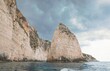 © Damian - Majestic cliffs rise from the azure sea, showcasing Greece's stunning landscapes. A boat glides through the water, offering breathtaking views of the rocky formations against a cloudy sky