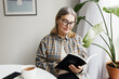 © shurkin_son - Pretty lady of 60s in casual clothes and glasses enjoying reading in morning, sitting next to windowsill with big houseplant in pot at white table with cup of tea and smartphone, holding book in hands
