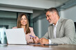 © insta_photos - Two professional middle aged business man and woman managers in office. 2 smiling executives using laptop discussing work. Busy colleagues talking on project strategy at meeting table. Authentic photo