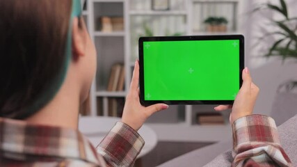 Closeup shot of girl sitting on sofa using tablet in horizontal position with green screen mockup in bright room using different gestures while watching video. Over the shoulder view.