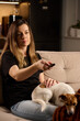 © Lazy_Bear - Woman sitting on sofa holding TV remote control. Female watching television at home with pet dog on couch. Concept of relaxation, lifestyle, and domestic life