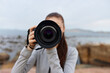 © SHOTPRIME STUDIO - Young female photographer capturing nature, holding a camera, wearing casual gray outfit, with a serene coastal background and soft, muted colors