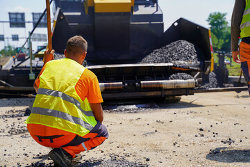 Fototapete - Construction worker supervising asphalt paver to ensure perfect application of fresh asphalt layer on highway construction site