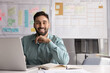 © fizkes - Happy confident young Latin businessman posing at office workplace, sitting at shared table with laptop alone, looking at camera, touching chin, smiling, laughing. Male business professional portrait