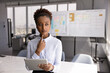 © fizkes - Serious young African American businesswoman holding tablet, thinking on online project electronic report, standing with digital gadget in office alone, looking away, touching chin, making decision