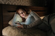 © dikushin - Attractive young woman lying in bed at night, using mobile phone smiling looking to screen, enjoying leisure time in cozy bedroom, illuminated by cell screen. Concept of mobile addiction and insomnia