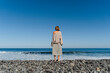 © Westend61 - Woman standing on a pebble beach looking at the ocean under a clear blue sky