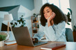© deagreez - Young woman with curly hair seated at a desk with a laptop, showing contemplative expression in a cozy home office setting