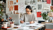 © Sergey - Asian female designer working at a desk with sketches and a laptop, surrounded by creative inspiration materials in a well-lit studio.
