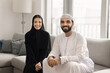© fizkes - Portrait of happy Arabic family posing for camera seated on cozy couch, young beautiful woman dressed in black abaya, her husband wearing white traditional kandura spend time together at modern home