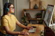 © TrueFrame Collective - Side view shot of cheerful young Caucasian woman in headphones sitting at desktop computer, writing prompt to generate image online