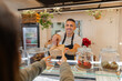 © BASILICOSTUDIO STOCK - Smiling man behind the counter giving a takeout box to a customer. Cozy small business with plants, jars, and friendly vibes. Ideal for takeaway or hospitality concepts. Horizontal medium shot.
