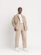 © GATE 22 - Young woman standing in a studio with a plain white background. she is wearing a beige blazer with a high collar and long sleeves, paired with beige trousers and white sneakers.