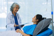 © ArLawKa - An elderly Asian female doctor is writing on a clipboard with a young female patient in a hospital room.