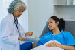 © ArLawKa - An elderly Asian female doctor is writing on a clipboard with a young female patient in a hospital room.