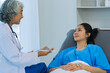 © ArLawKa - An elderly Asian female doctor is writing on a clipboard with a young female patient in a hospital room.