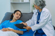 © ArLawKa - An elderly Asian female doctor is writing on a clipboard with a young female patient in a hospital room.