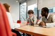 © Drobot Dean - A woman in her 30s sitting at a table in the office with a young White man and an adult Black man during a meeting in the conference room with big windows with a city view with skyscrapers
