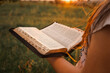 © shine.graphics - Close-up of a girl reading the Bible outdoors during golden hour. The soft sunset light gently illuminates the open pages, symbolizing faith, peace, and personal devotion