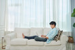© Dialogue Frame - A young Asian man wearing a blue shirt and jeans is sitting on the living room sofa, working on a laptop.
