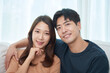 © Dialogue Frame - Young Asian man and woman sitting on a living room couch, affectionately putting their arms around each other's shoulders