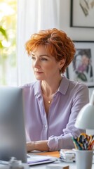 Fototapete - Middle-aged woman working on computer in bright home office with natural light, wearing purple shirt and gold necklace