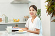 © Dialogue Frame - Young Asian woman in a white T-shirt sitting at a kitchen dining table chair, reading while eating bread and drinking milk