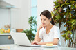 © Dialogue Frame - Young Asian woman in a white T-shirt sitting at a kitchen dining table chair, working on a laptop while eating bread and drinking milk