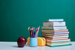 © Татьяна Кутина - Stack of colorful books, a mug filled with colored pencils, an apple, and a yellow pencil case are arranged on a desk against a green chalkboard background, depicting academic theme.