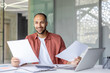 © Liubomir - Successful businessman doing paperwork, man in casual clothes checking documents and bills, satisfied with results of activity smiling, working inside office with laptop