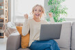 © deagreez - Excited blonde woman sitting on a comfortable sofa using a laptop, expressing happiness and success in a cozy home interior