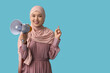 © Pixel-Shot - Young Asian Muslim woman with megaphone pointing at something on blue background. National Be Heard Day