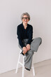 © Viktor Solomin/Stocksy - Mature woman with gray hair sits confidently on a stool