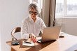 © Viktor Solomin/Stocksy - Senior woman working on laptop at home during the day with notepad
