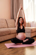 © dvulikaia - Beautiful young pregnant brunette woman is sitting in a lotus position and practising kundalini yoga in the living room opposite the sofa
