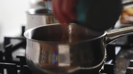 A person is pouring something into a pot on a stove. The pot is silver and has a handle