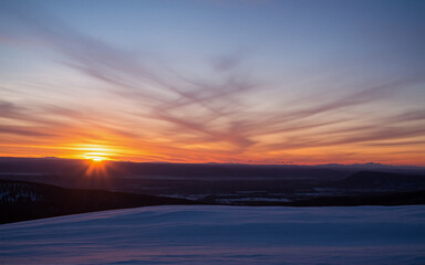 Aufkleber - A breathtaking sunset paints the sky with warm orange and cool blue hues, casting a serene glow over a snowy, mountainous landscape in the distance.