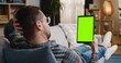 © VAKSMANV - Static back view of Caucasian male with glasses holding tablet device vertically with chroma key on screen. Laying on sofa in apartment and resting. Wearing white sneakers, jeans and checkered T-shirt