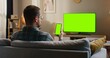 © VAKSMANV - Back view of Caucasian man sitting on couch in front of large television with green screen. Holding smartphone vertically with one hand. Mobile device with chroma key on screen. Looking with focus.