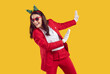 © Studio Romantic - Studio shot of cheerful brunette European woman wearing red Santa Claus suit, sunglasses and funy firtree horns, dancing carefree against vivid yellow background, has festive mood