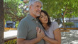 © Krakenimages.com - Middle-aged couple embracing outdoors in a sunny urban park, expressing love and togetherness, with man and woman standing closely amidst greenery and modern urban landscape.