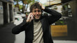 © Krakenimages.com - Frustrated man talking on phone outdoors in city wearing black jacket and beige sweater, hair disheveled, standing near street with blurred background, expressing stress and urgency