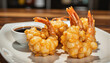 © f_bossa - Modern food photo advertisement: Crispy coconut shrimp with tails attached rests on a white square plate next to a small bowl of dark sauce.