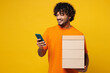 © ViDi Studio - Young fun happy Indian man he wear orange t-shirt casual clothes hold stack cardboard blank boxes use mobile cell phone online isolated on plain yellow background studio portrait. Lifestyle concept.