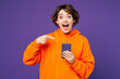 © ViDi Studio - Young surprised happy Caucasian woman wear orange hoody casual clothes hold in hand use point index finger on mobile cell phone isolated on plain purple background studio portrait. Lifestyle concept.