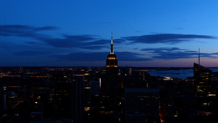 Poster - A tall skyscraper brightly illuminates the dark cityscape below under a twilight sky. Evening descends over the urban sprawl in a scenic panoramic view.